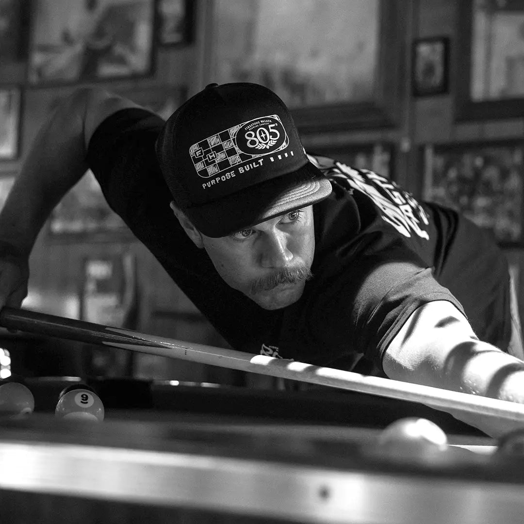 Person wearing 805 Purpose Black Hat, Man leaning over pool table in indoor garage setting - Slowride Supply