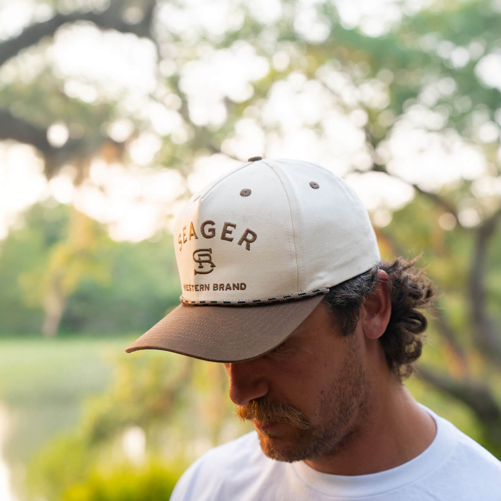 Person wearing Branded Snapback Cream/brown Hat, Garden setting with flowering tree branches and soft green foliage in