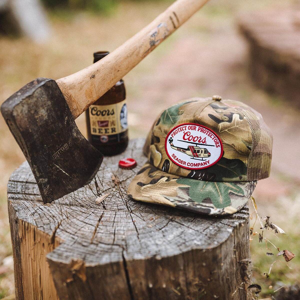 Person wearing Coors Banquet Helo Trucker Green Hat, Weathered tree stump with axe and beer bottle in outdoor work setting -