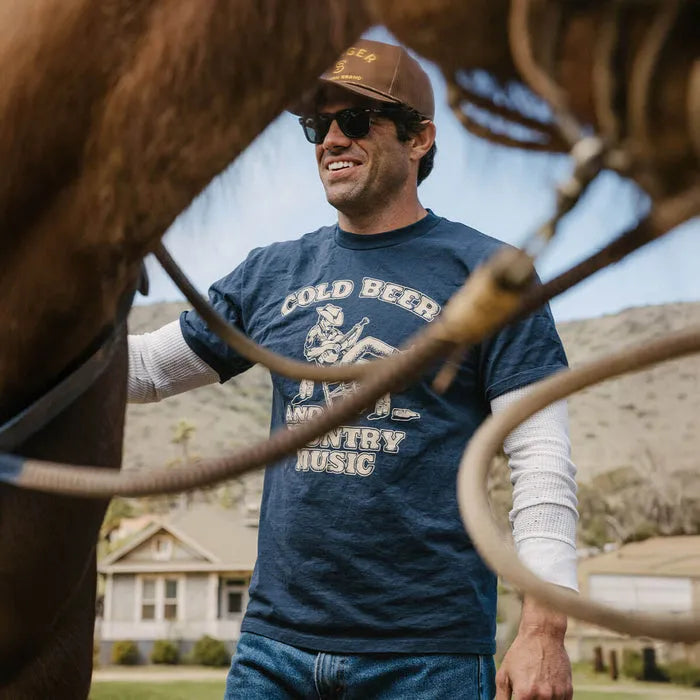 Person wearing Country Music Navy Tee, Man wearing navy tee standing outdoors near wooden ranch fence with desert landscape