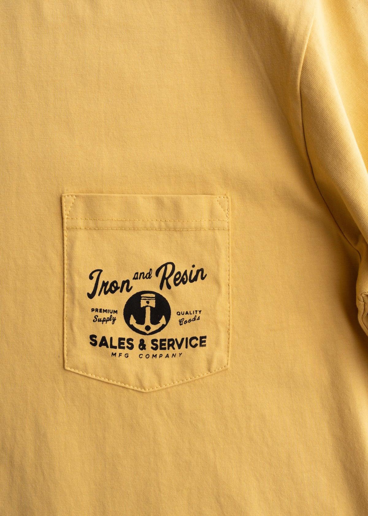 Close-up of Old Cowboy Pocket Black Tee showing Yellow work shirt with chest pocket featuring black anchor graphic and "Iron