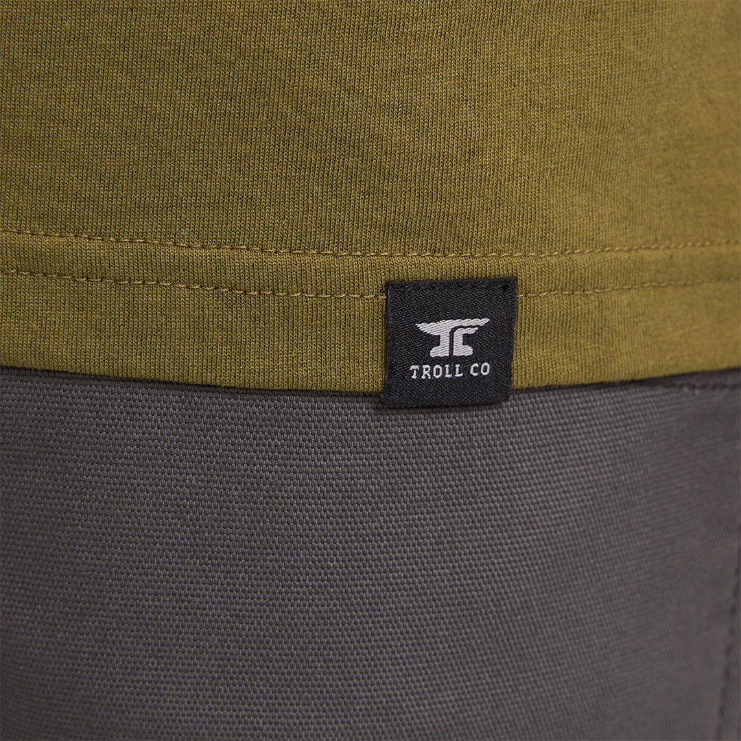 Close-up of Pay Me Military Tee showing Black woven label with white anvil logo and "TROLL CO" text on olive and charcoal