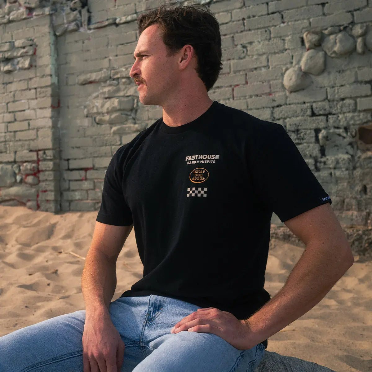 Person wearing Pursuit Natural Tee, Man sitting against weathered brick wall in industrial setting - Slowride Supply