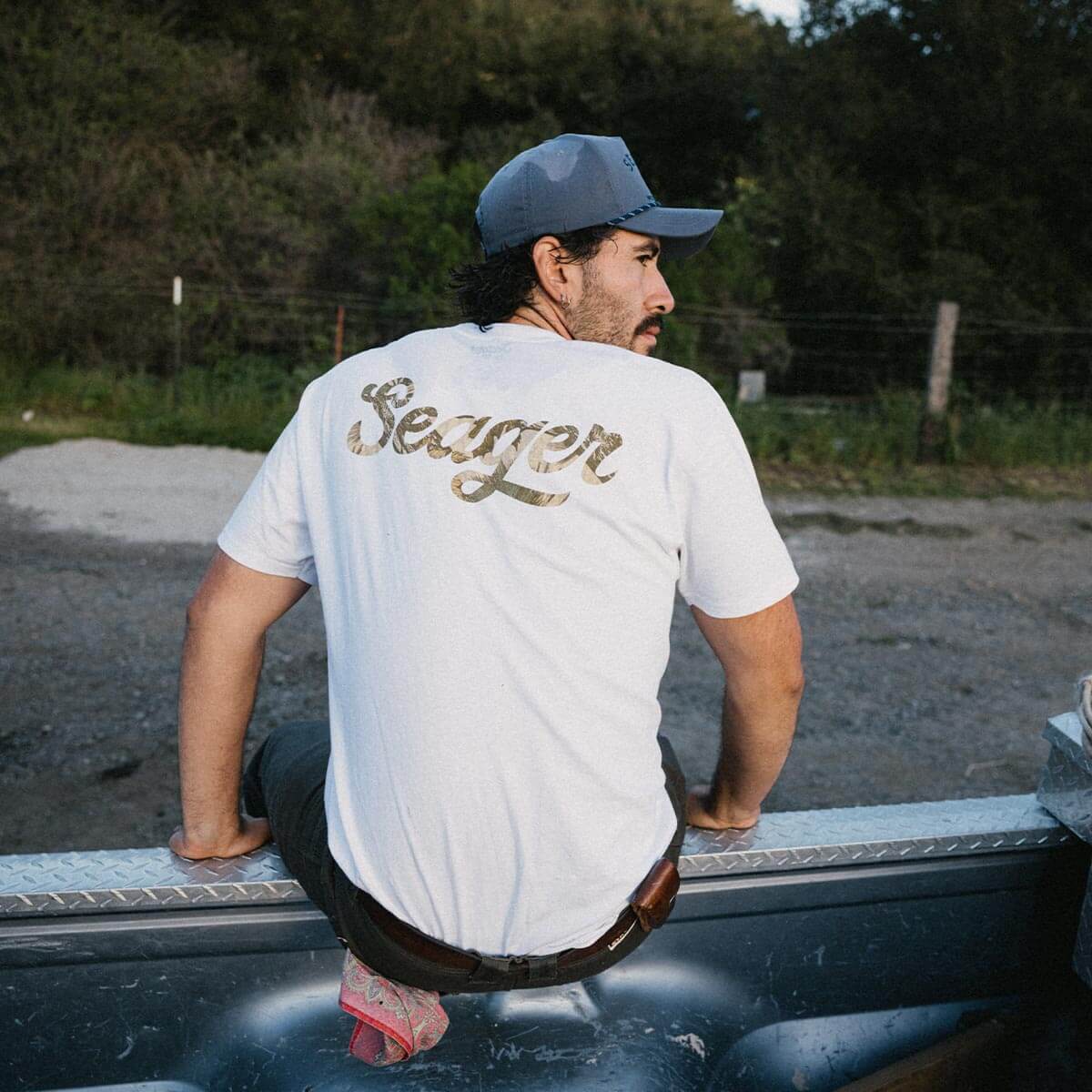 Person wearing Russ Fill Black Tee, Man seated on truck bed in wooded area with evergreen trees behind - Slowride Supply