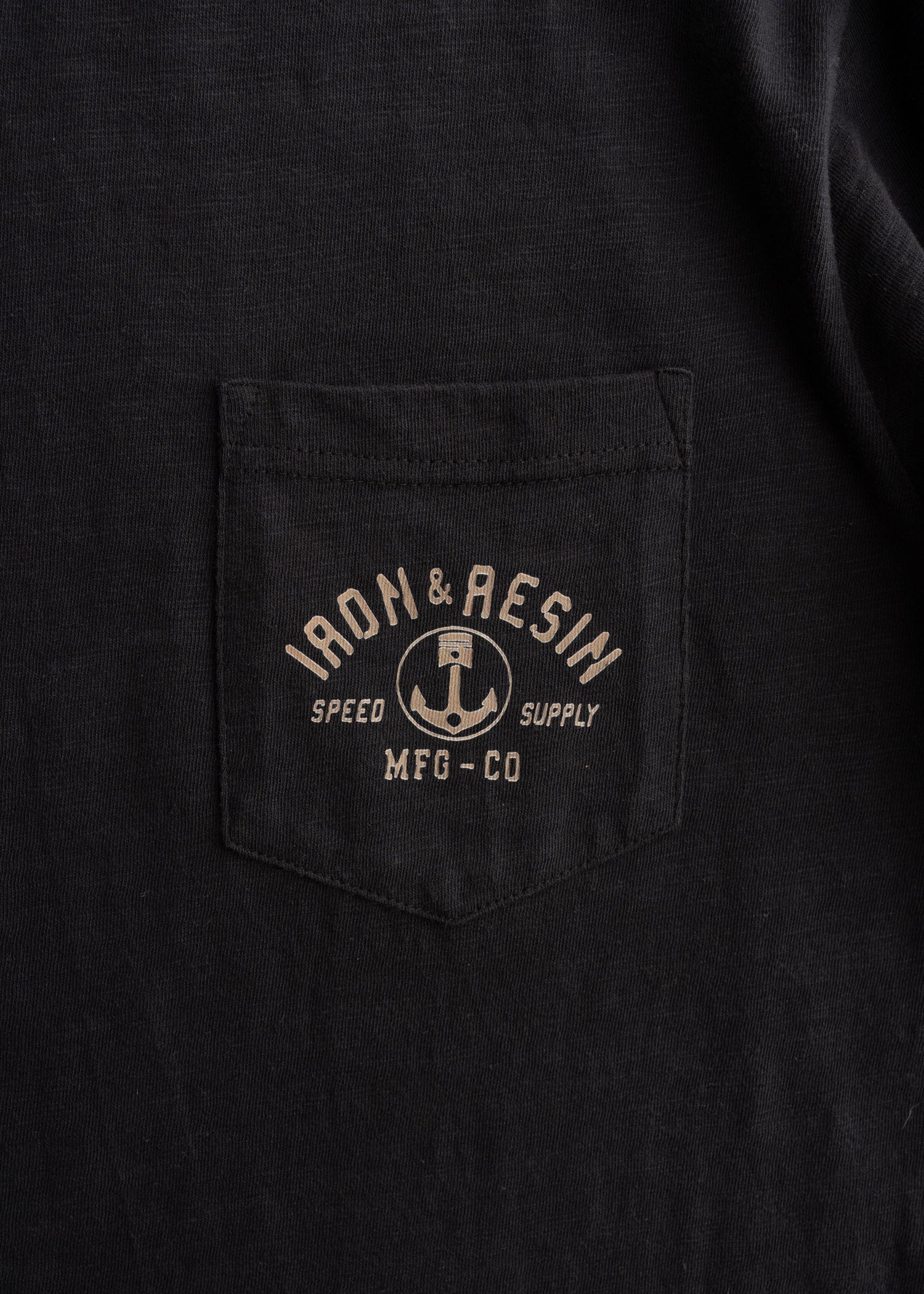 Close-up of Speed Supply Black Tee showing Chest pocket with tan anchor graphic and "Iron & Resin Speed Supply Mfg-Co" text