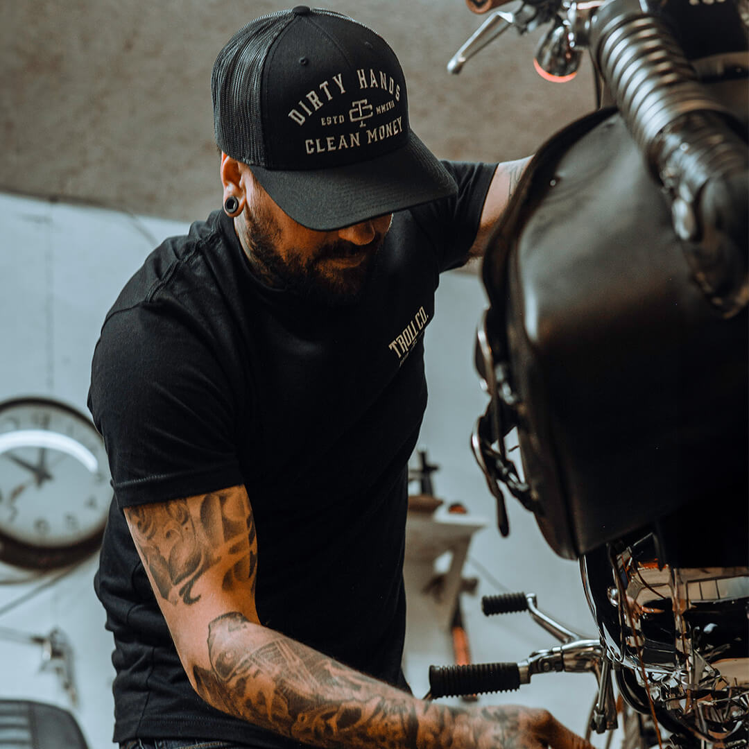 Person wearing DHCM Classic Trucker White/coyote Brown Hat, Person working on motorcycle engine in industrial garage setting