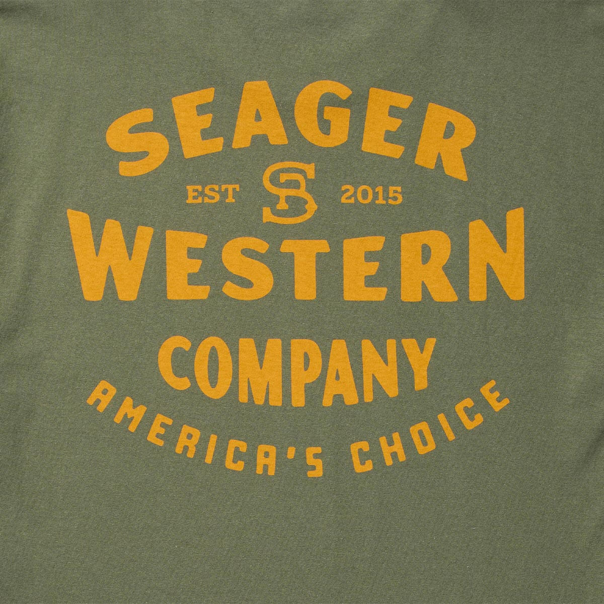 Close-up of Americas Choice Surplus Green Tee showing Gold block lettering and monogram emblem on surplus green cotton