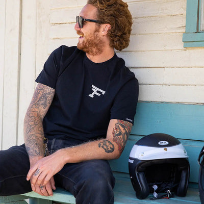 Person wearing Arthur Black Tee, Man seated against weathered white and turquoise painted wooden wall next to racing helmet