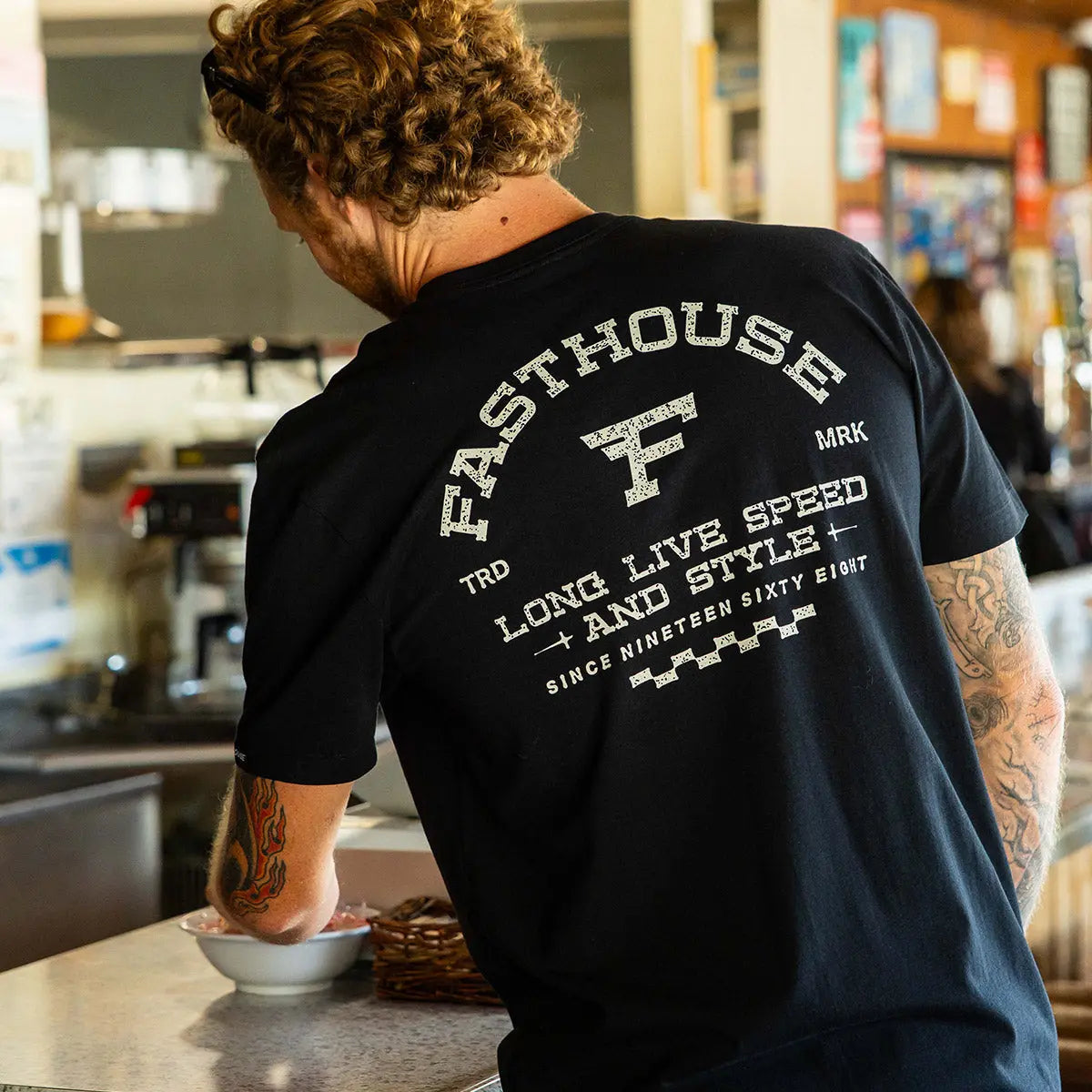 Person wearing Arthur Black Tee, Indoor kitchen setting with countertop and shelving in background - Slowride Supply