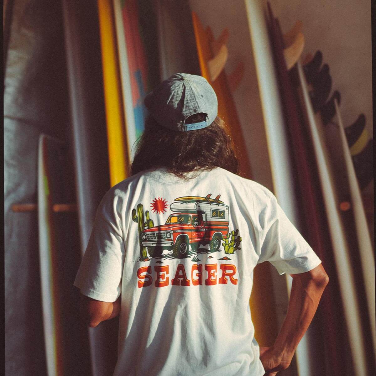 Person wearing Baja Vintage Black Tee, Desert backdrop with colorful surfboards and warm golden light casting long shadows -