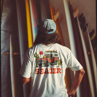 Person wearing Baja Vintage Black Tee, Desert backdrop with colorful surfboards and warm golden light casting long shadows -