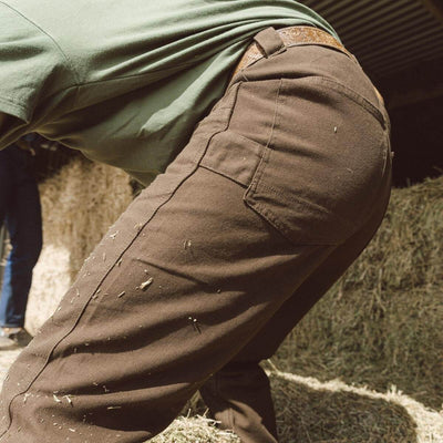 Person wearing Bison Canvas Pants Brown Pants, Worn against a weathered garage wall with concrete blocks and straw bedding