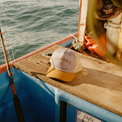 Person wearing Branded Snapback Cream/brown Hat, Cream and yellow snapback resting on weathered wooden boat deck overlooking
