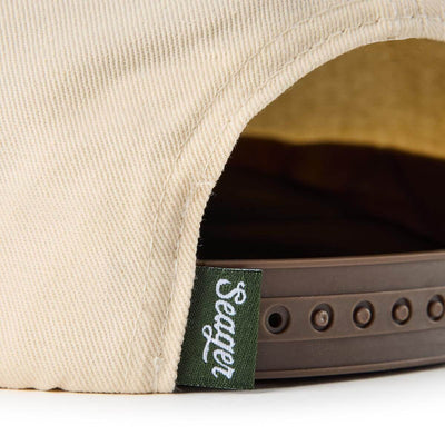 Close-up of Branded Snapback Hat Apparel Cream/Camo Apparel showing Green woven label with white script text on snapback hat