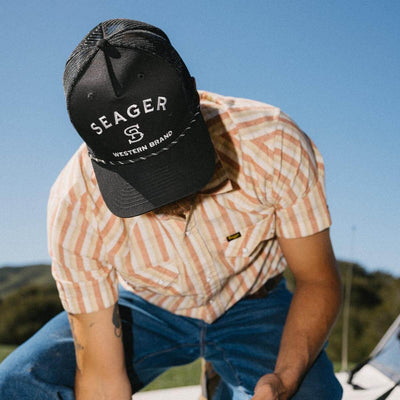Person wearing Branded Trucker Black/Black Hat, Person wearing black trucker hat outdoors in open desert landscape under