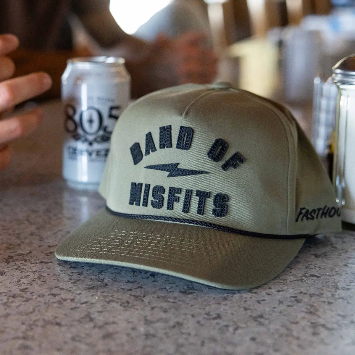 Person wearing Capone Olive Hat, Tan cap resting on concrete bar counter with beer cans nearby - Slowride Supply