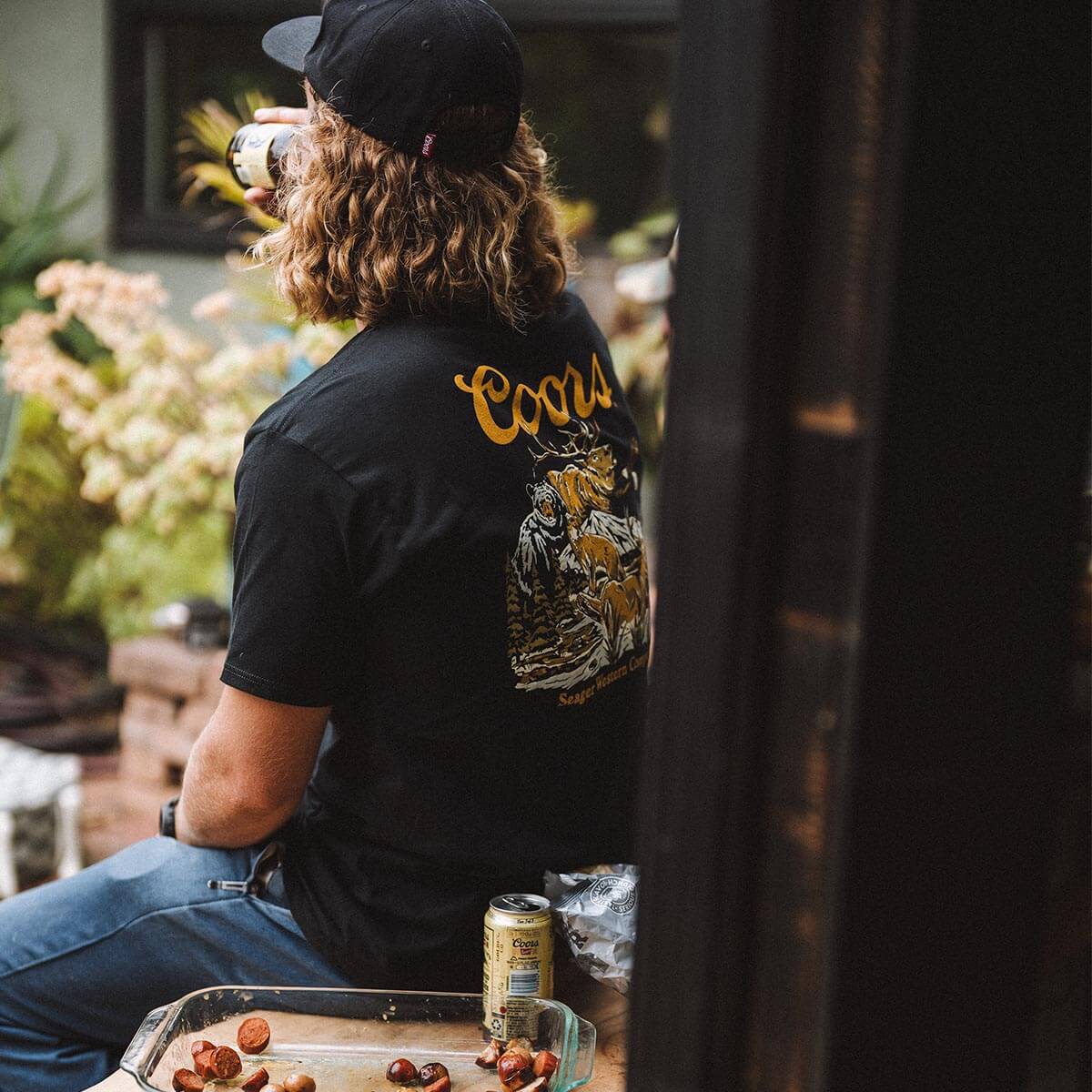 Person wearing Coors Banquet Preserve Black Tee, Worn in a backyard garden setting with flowering plants and wooden