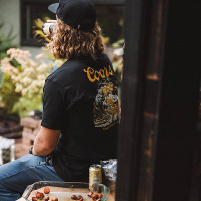 Person wearing Coors Banquet Preserve Black Tee, Worn in a backyard garden setting with flowering plants and wooden