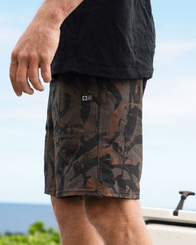 Person wearing Crew Boatshort 18" Boardshorts Camo Boardshorts, Waterfront setting with clear sky and boat visible in