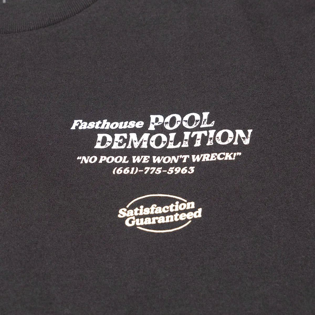 Close-up of Demolition Black Tee showing White screen-printed text and badge on charcoal ribbed cotton fabric, phone number