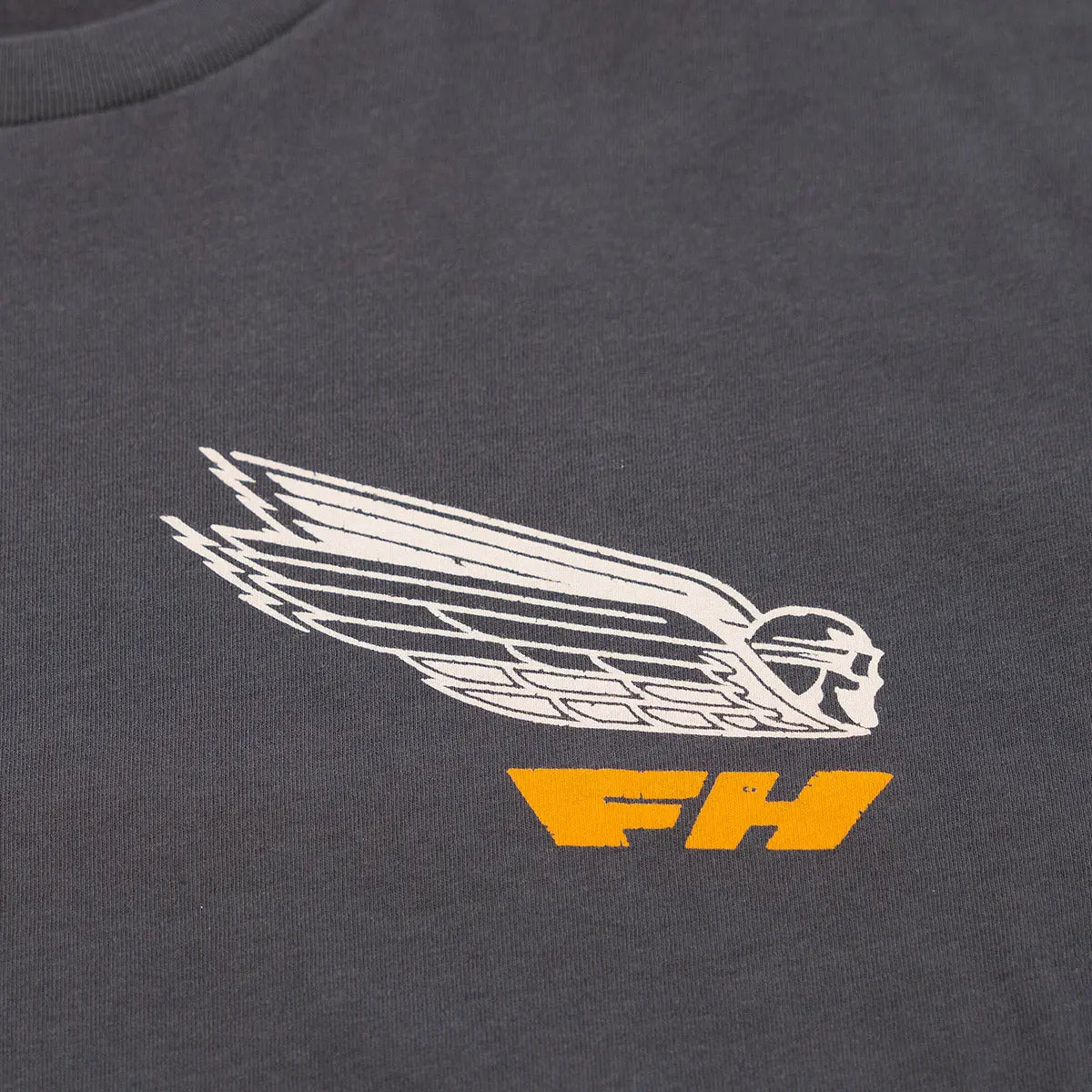 Close-up of Flyer White Tee showing Embroidered winged figure in cream with gold lettering below on charcoal fabric -