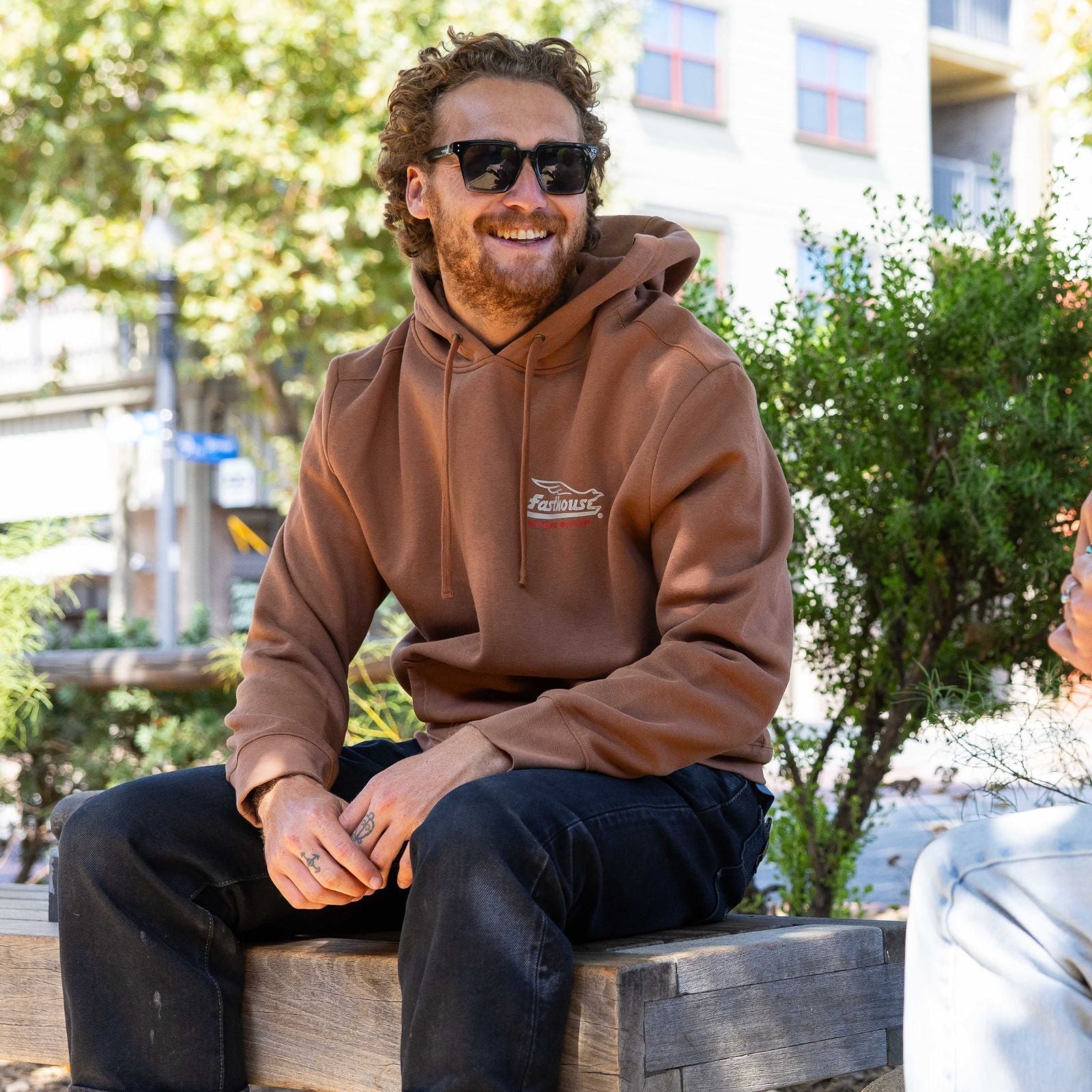 Person wearing Gilmore Hooded Pullover Apparel Chestnut Apparel, Urban courtyard with green foliage and white building