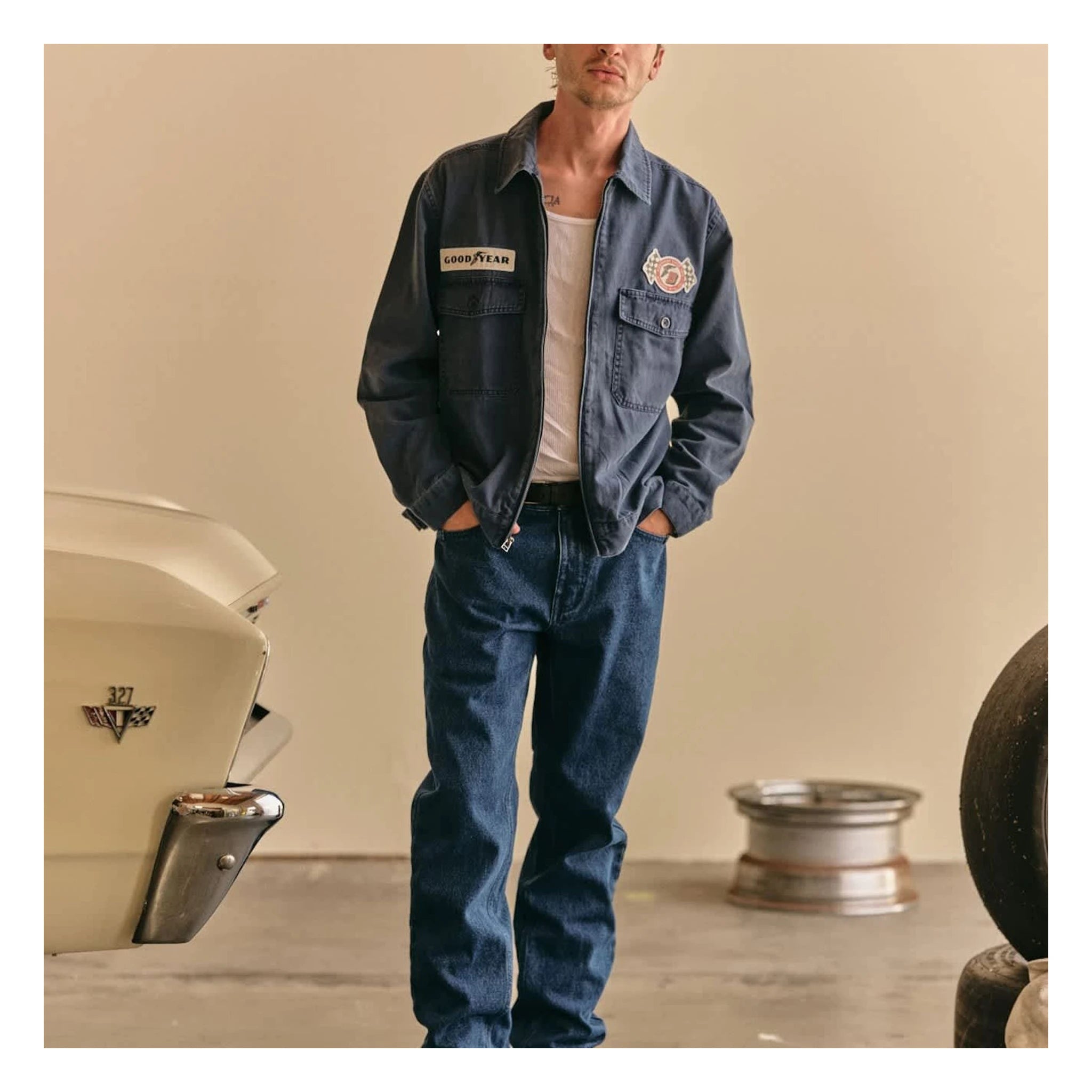 Person wearing Goodyear Trademark LW Washed Navy Jacket, Garage setting with vintage car and racing wheels visible -