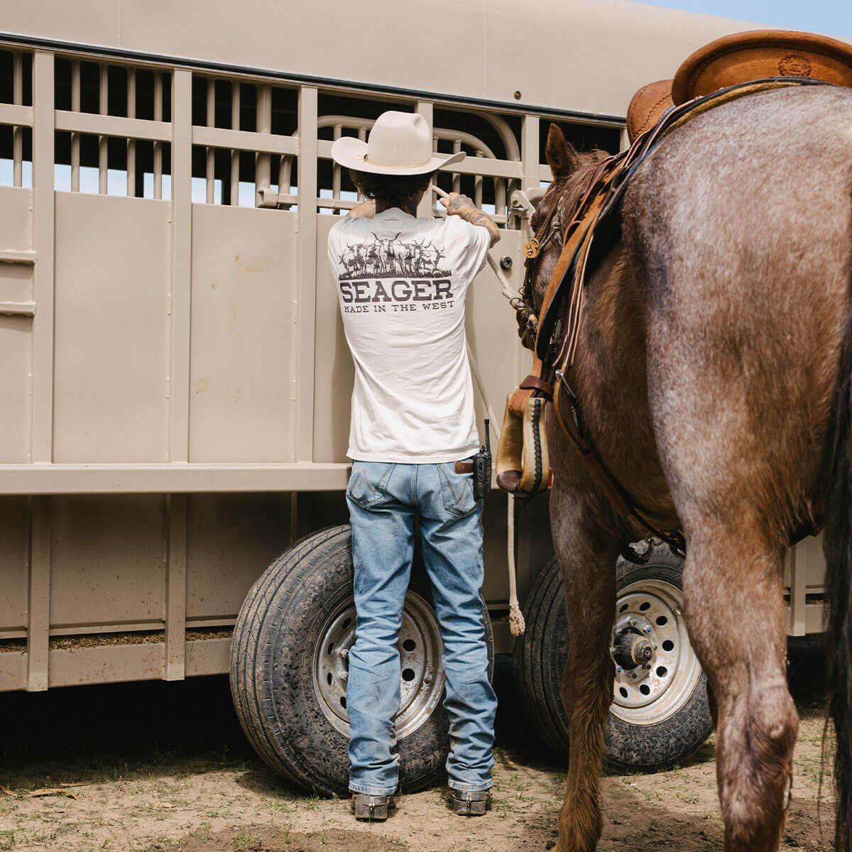 Person wearing Herd Vintage White Tee, Cowboy in white tee standing at horse trailer in ranch setting - Slowride Supply