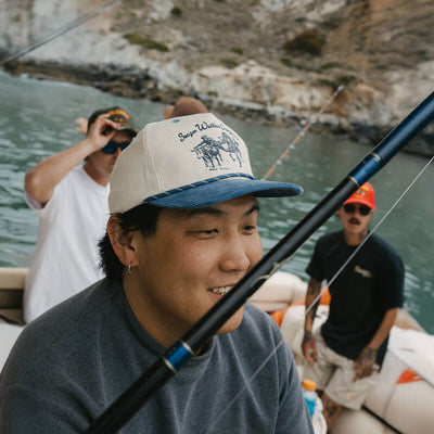Person wearing Los Rios Cordouroy Snapback Cream/green Hat, Boat on water with rocky cliffs and fishing gear visible -