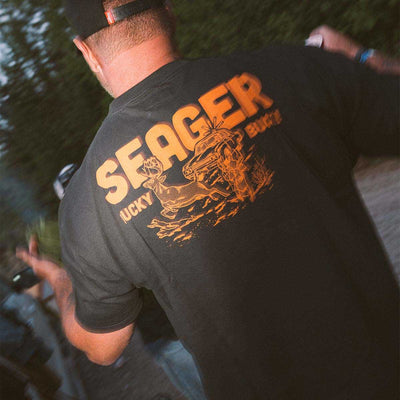 Person wearing Lucky Buck Black Tee, Person wearing black tee near water and greenery outdoors - Slowride Supply