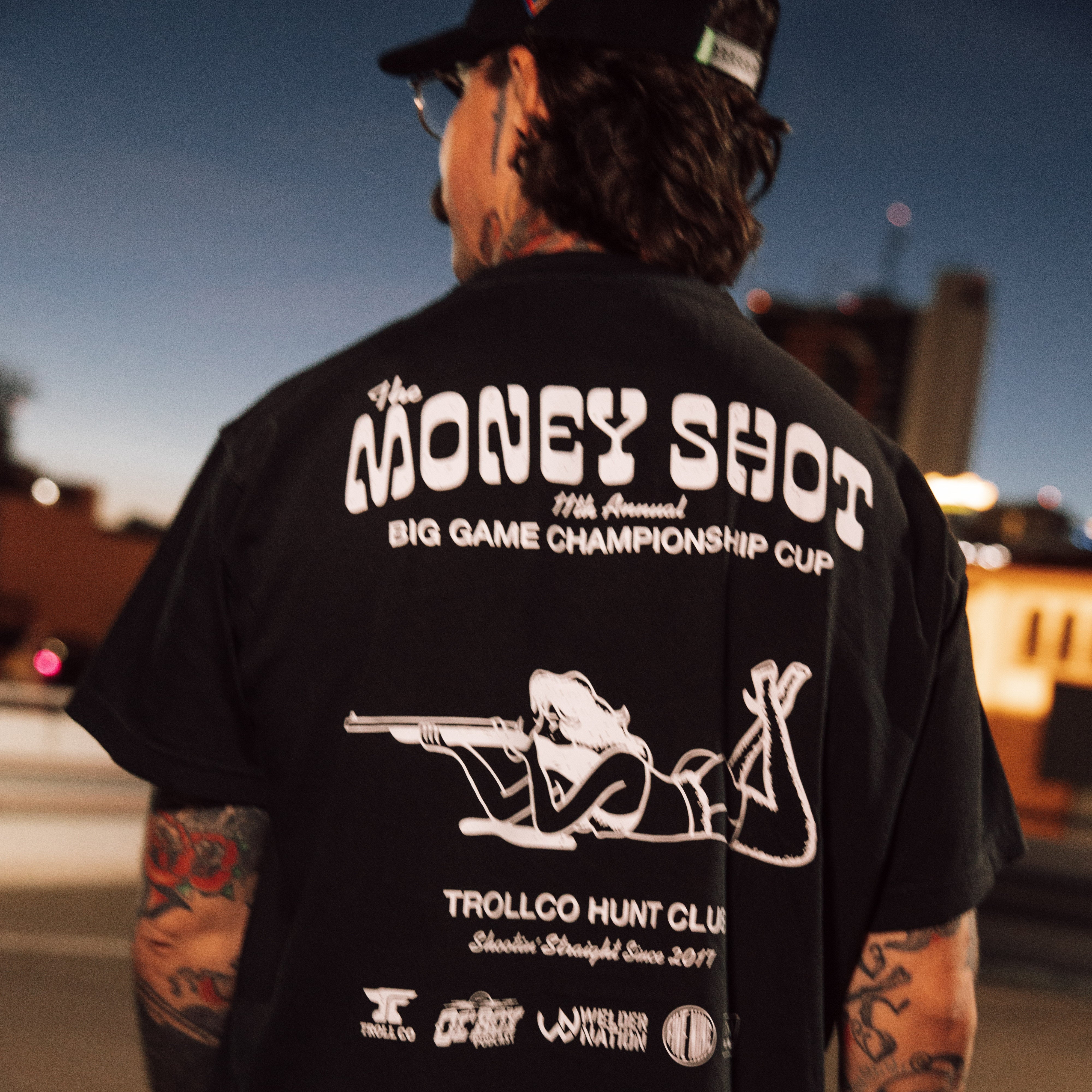 Person wearing Money Shot Heavyweight Tee, Urban street at dusk with industrial buildings and neon lights - Slowride Supply