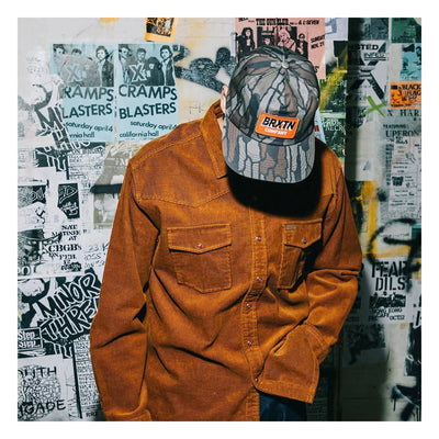 Person wearing New West Relaxed Overshirt Washed Copper Cord Jacket, Worn against a wall covered with layered punk and