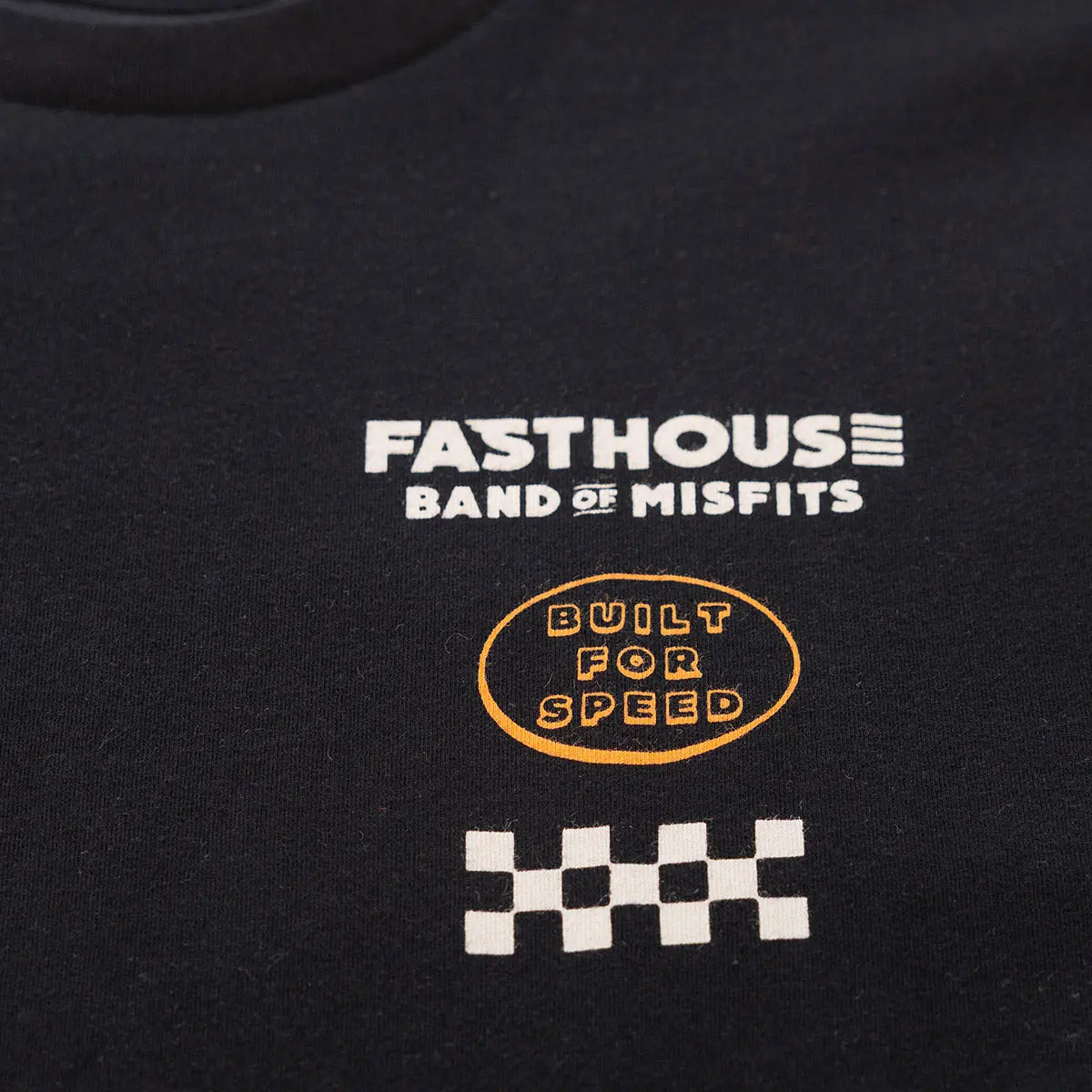 Close-up of Pursuit Natural Tee showing White text and gold oval badge with checkered flag graphic on black fabric -
