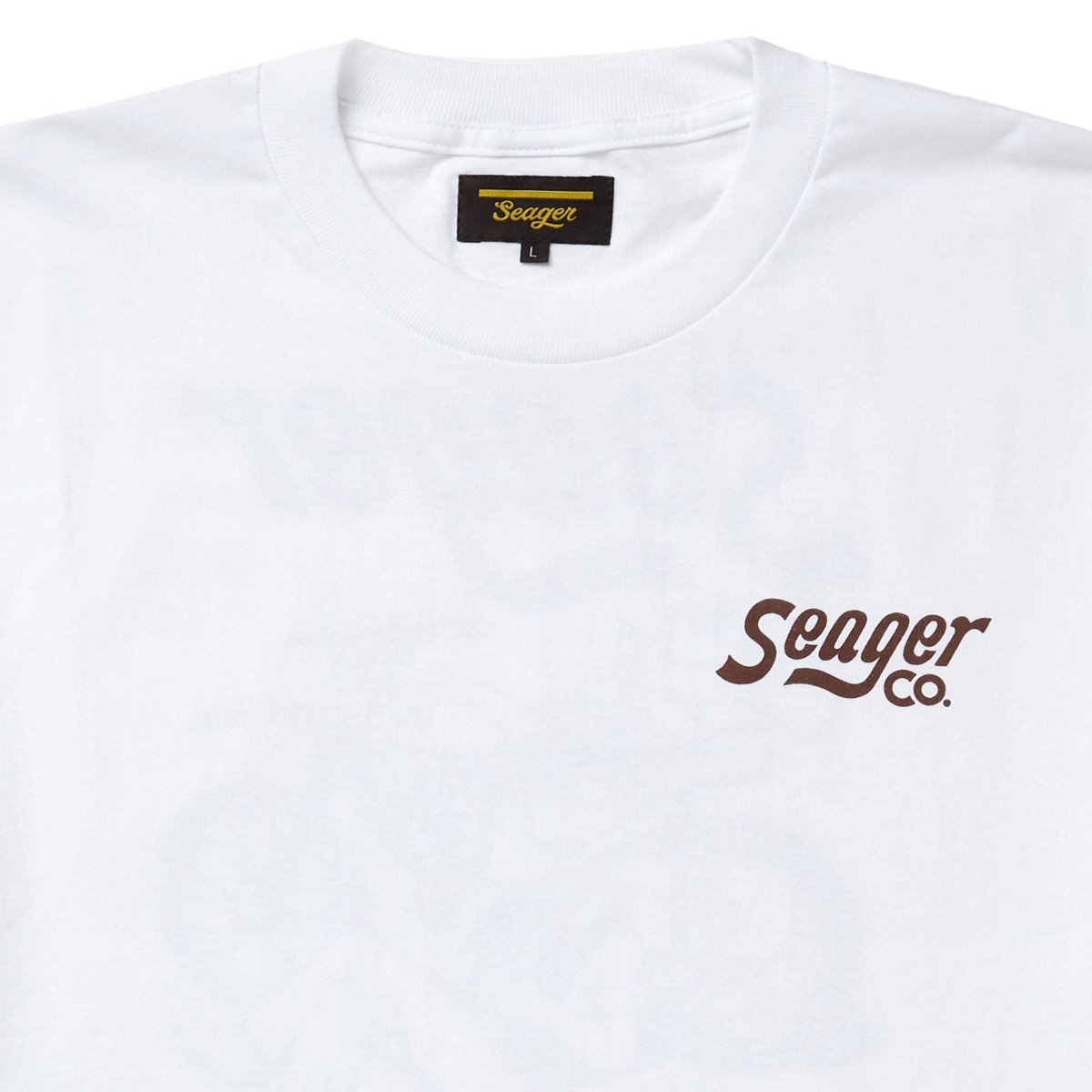 Close-up of Romero White Tee showing White with brown script logo on chest, black woven label at neckline with gold text -