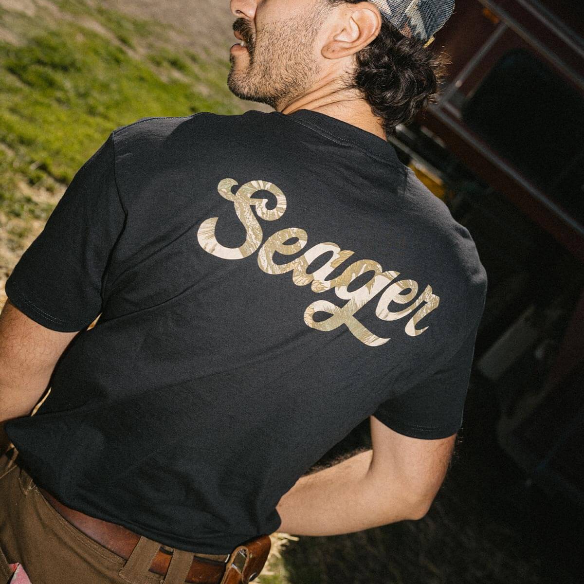 Person wearing Russ Fill Black Tee, Man wearing black tee outdoors near moss-covered stone wall and garage entrance -