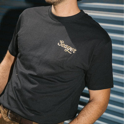 Person wearing Russ Fill Black Tee, Man wearing black tee standing against corrugated metal wall in daylight - Slowride