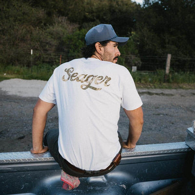 Person wearing Russ Fill Black Tee, Man seated on truck bed in wooded area with evergreen trees behind - Slowride Supply