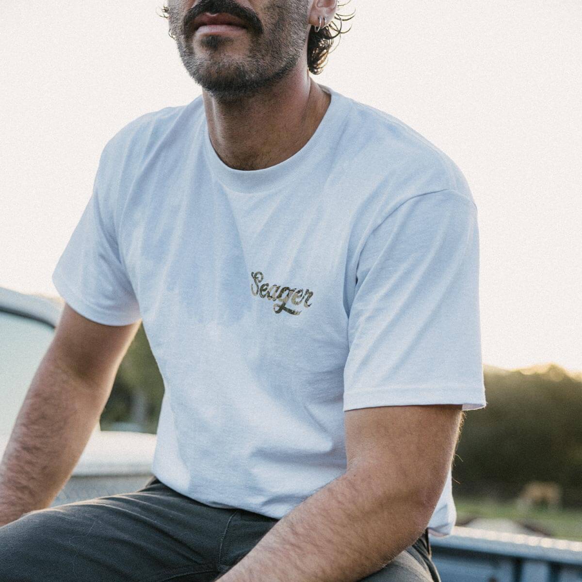Person wearing Russ Fill Black Tee, Man seated outdoors near water with blurred landscape background - Slowride Supply