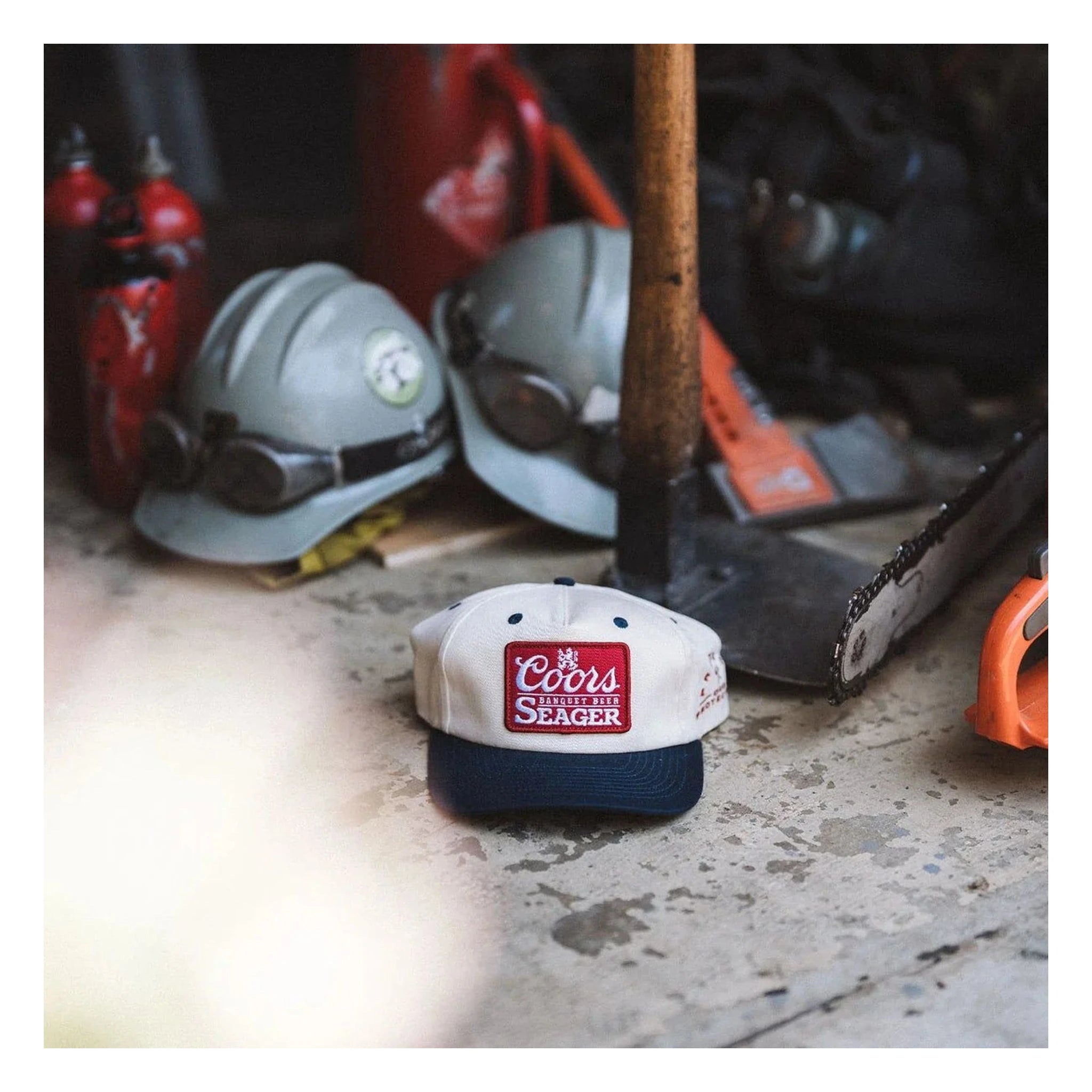 Person wearing X Coors Banquet P.o.p. Snapback Cream/navy-cream/navy Hat, Garage floor surrounded by racing helmets, tools,