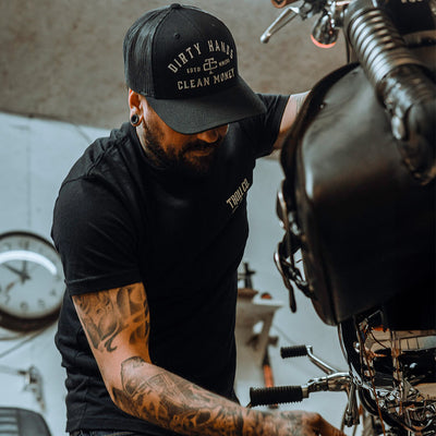Person wearing DHCM Classic Trucker White/coyote Brown Hat, Person working on motorcycle engine in industrial garage setting