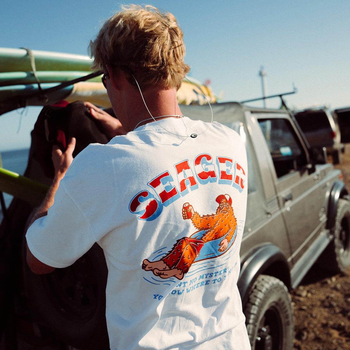 Person wearing Tubular White Tee, Beach parking lot with vintage trucks and clear sky in background - Slowride Supply