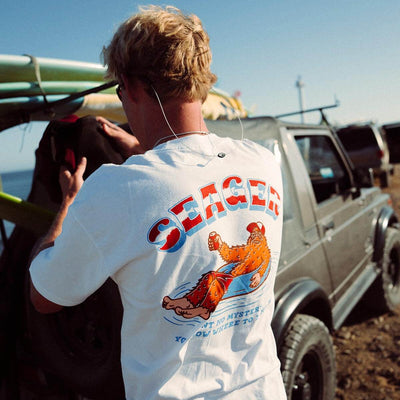 Person wearing Tubular White Tee, Beach parking lot with vintage trucks and clear sky in background - Slowride Supply