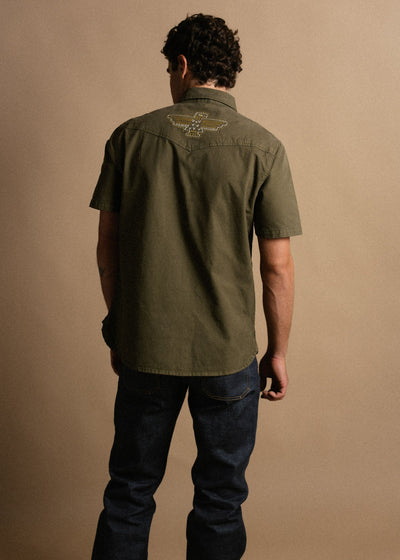 Person wearing Westy Shirt S/S Woven Light Khaki S/S Woven, Neutral studio setting with tan backdrop, model standing against